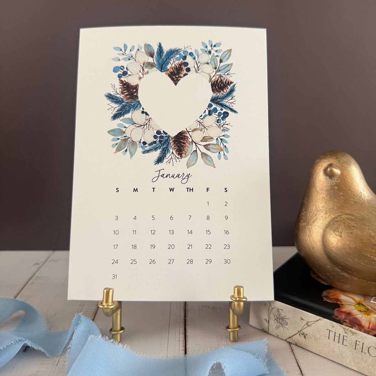 January page of the 2027 desk calendar with a winter wreath of blue foliage and pinecones